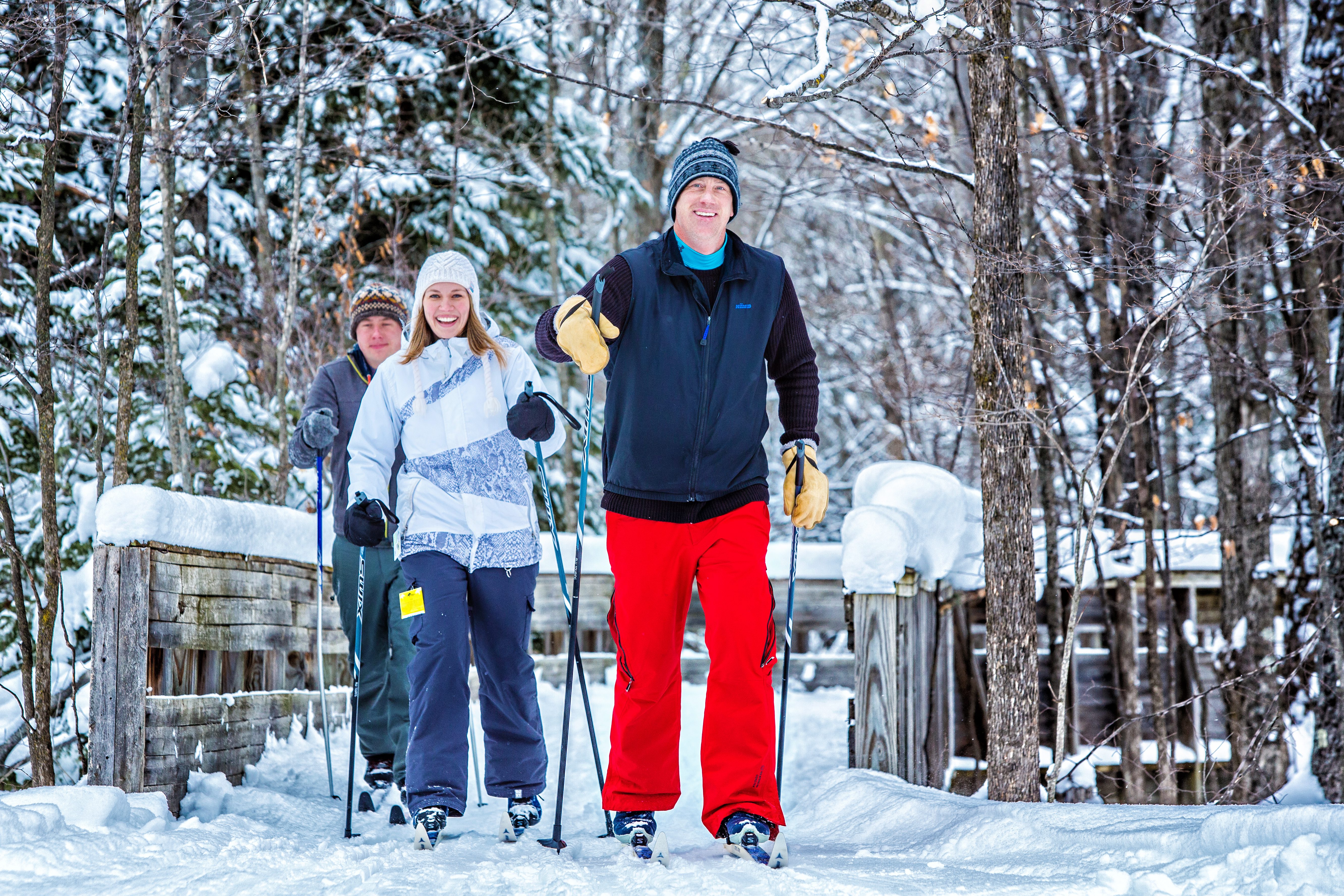 Three adults cross country skiing across a bridge on a sunny, snowy day at Treetops Resort.