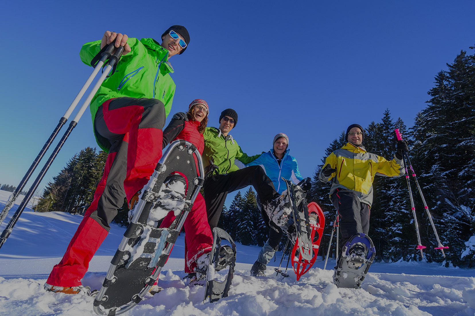 A group of people in bright winter gear raising their snow shoes up from the snow.