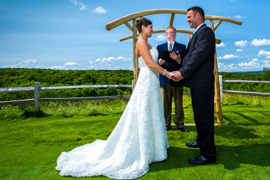 A bride, groom, and officiant stand at an arch in front of a farm fence on a bright, sunny day.