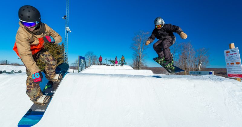Two snowboarders hit the jumps on a sunny day at Treetops Resort.