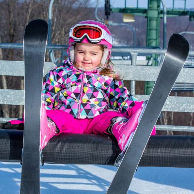 Young girl in pink snowsuit and goggles on a chairlift wearing skis